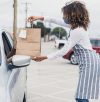 A young adult waitress wears her protective mask to deliver a curbside order to an unrecognizable person in a car.  The waitress stands away from the car.